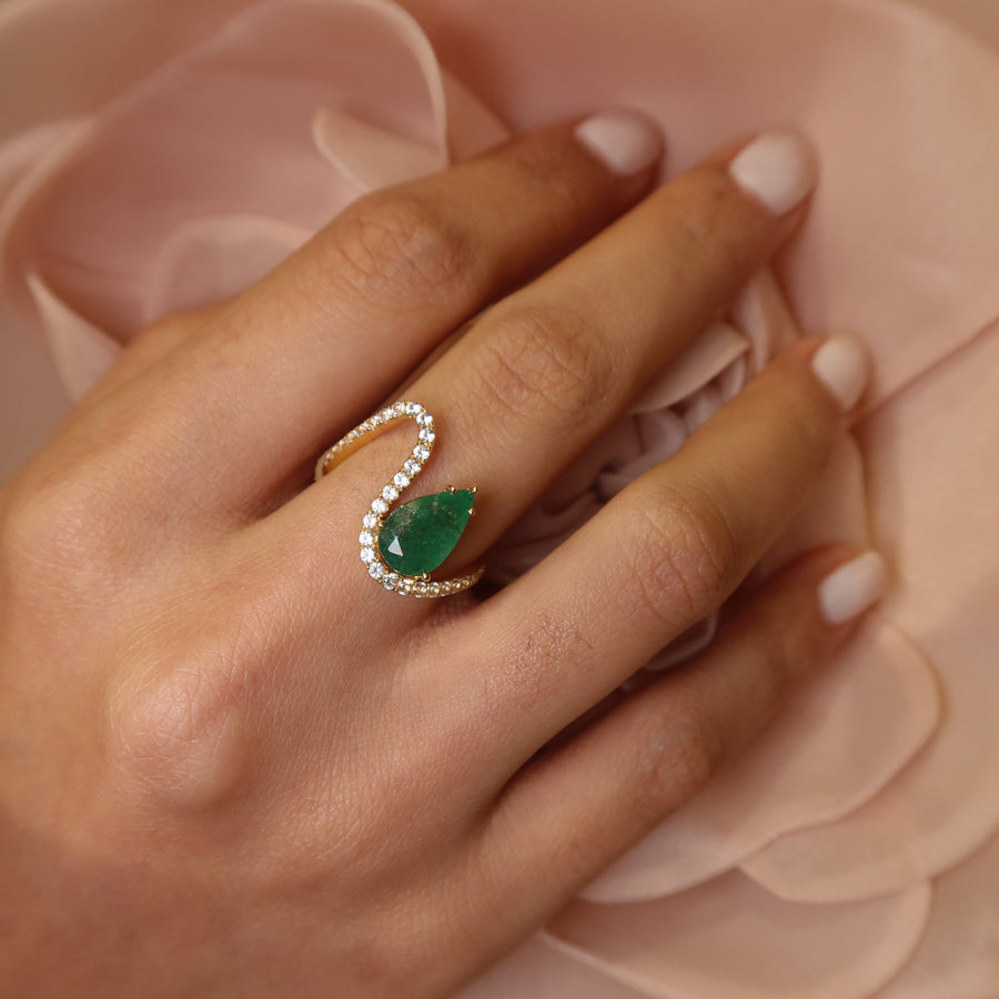 Hand wearing a 18k yellow gold zambian emerald and white sapphires gemstone ring on a soft pink fabric background