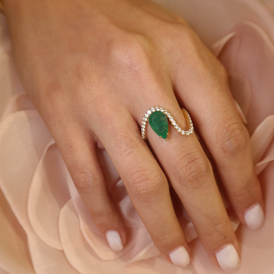 Hand wearing a 18k yellow gold zambian emerald and white sapphires gemstone ring on a soft pink fabric background