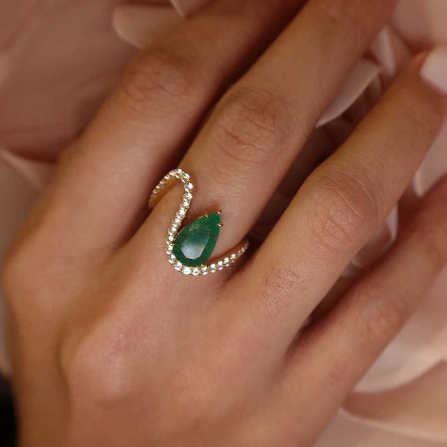 Hand wearing a 18k yellow gold zambian emerald and white sapphires gemstone ring on a soft pink fabric background